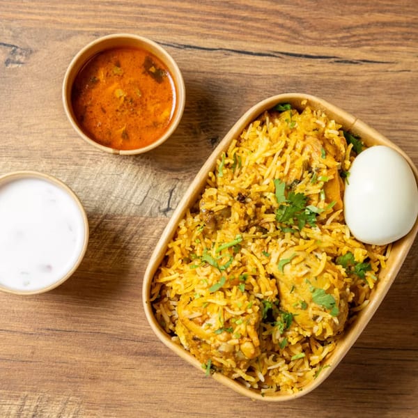 Chicken biryani with a boiled egg, a side of curry, and a side of raita on a wooden table.