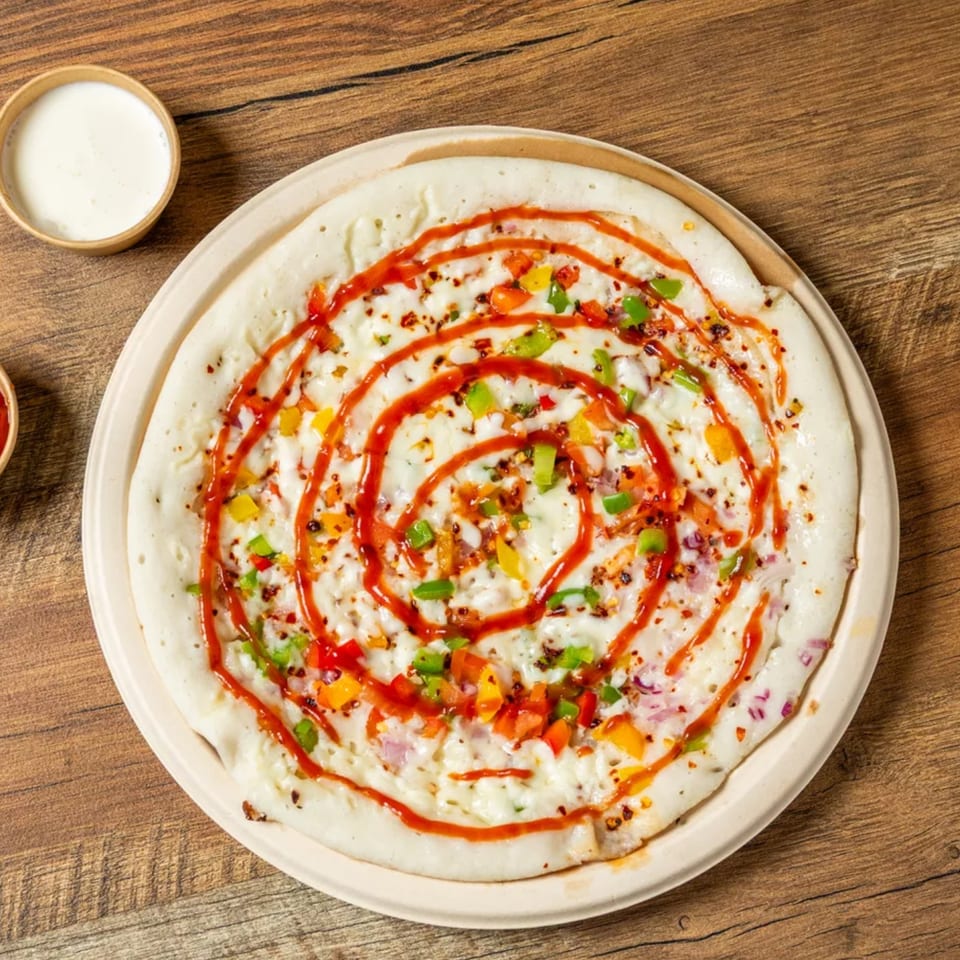 A white uttapam topped with colorful diced vegetables and a swirl of red sauce on a wooden table.