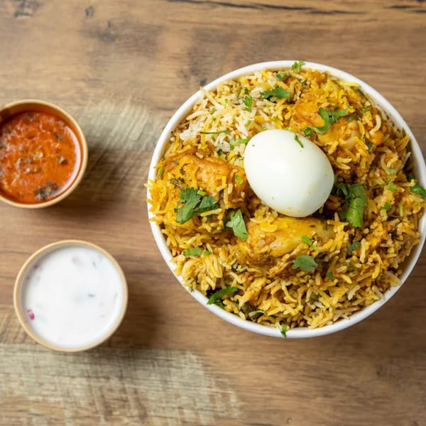 Overhead view of chicken biryani with a boiled egg, red sauce, and white raita on a wooden table.