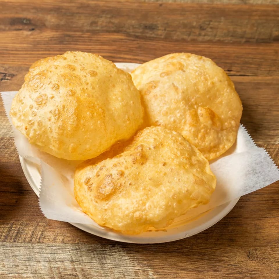 Three golden-brown, puffy puri breads on a white plate lined with parchment paper, on a wooden table.
